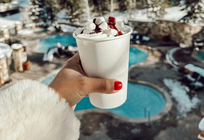 A selfie view of an outstretched hand holding a cup of hot chocolate covered in marshmallows and whipped cream at a ski spot.